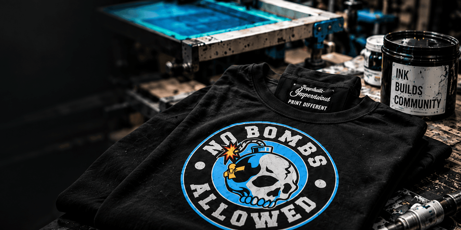 Black tee with a screen-printed No Bombs Allowed graphic resting on a press bed beside ink buckets